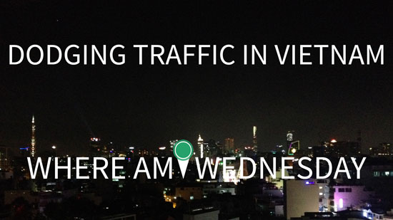 Photo of a city skyline at night with white text in the foreground that says "Dodging Traffic in Vietnam. Where Am I Wednesday"