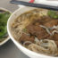 Photo contains a bowl of noodles with veg beef, mushrooms, sprouts, and other vegetables. On a separate plate, there are fresh herbs and leafy greens.
