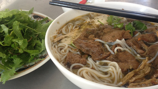 Photo contains a bowl of noodles with veg beef, mushrooms, sprouts, and other vegetables. On a separate plate, there are fresh herbs and leafy greens.