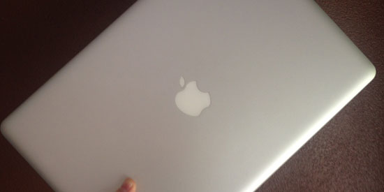 Image contains a grey MacBook Pro being held at an angle by a hand against a brown background.