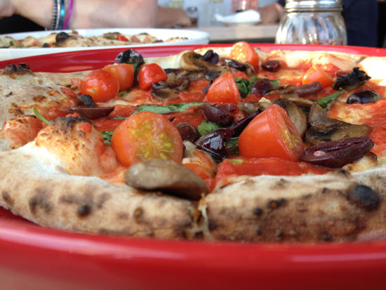 Image contains a wood-fired pizza with a slightly blackened crust and tomatoes, mushrooms, olives, and marinara sauce for toppings.