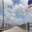 Image contains a wooden pier that narrows toward the centre of the image. Above it, there is a blue sky with white clouds. To the right, the Thailand flag can be seen with blue, white, and red stripes.
