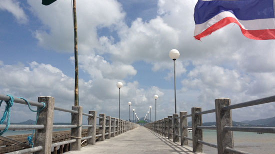 Image contains a wooden pier that narrows toward the centre of the image. Above it, there is a blue sky with white clouds. To the right, the Thailand flag can be seen with blue, white, and red stripes.