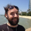 Image contains a smiling, bearded man with dark hair blowing in the wind. He's smiling and in the foreground while a sandy beach and palm trees can be seen in the background.