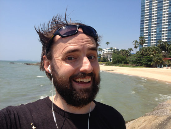Image contains a smiling, bearded man with dark hair blowing in the wind. He's smiling and in the foreground while a sandy beach and palm trees can be seen in the background.