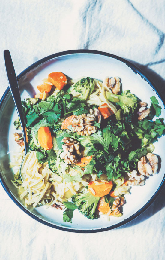 Image contains a pale blue plate with a dark blue rim on a light blue cloth. On the plate, there are a number of different vegetables along with noodles and a silver fork.