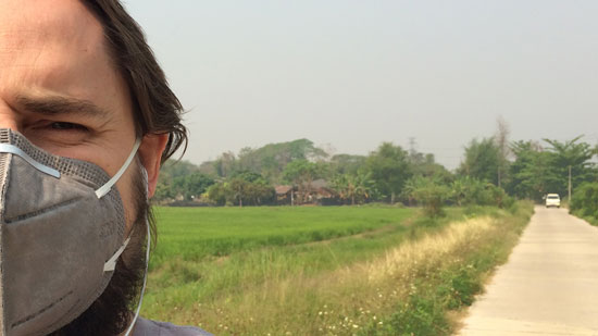 Image contains a photo of a man on the left-hand side wearing a face mask over his mouth and nose. To the right of the man, a hazy sky is visible above some trees in the horizon, a green rice field, and a narrow road.