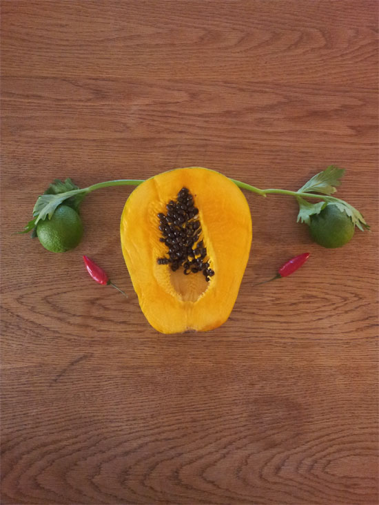 Image contains a medium-dark brown wooden background. On the wood, there is a variety of fruit and vegetables. In the centre, there is a papaya and coming from the left and right of the fruit are two small stalks of a leafy green vegetable. Nestled under the leaves at the end of each stalk are lemons, and just below each lemon on the right and left there is a small hot pepper. The whole image is meant to represent the female reproductive system.