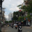 Image is a photo of a Saigon street with numerous scooters coming towards the camera. Shops are visible on both sides of the street, and a small neon sign in the top left-hand corner says "QuÃ¡n Chay GiÃ¡c Tha".