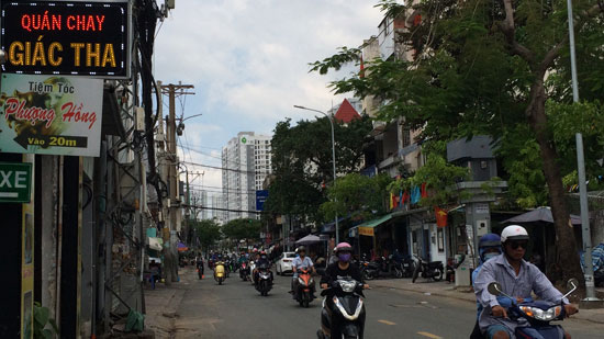 Image is a photo of a Saigon street with numerous scooters coming towards the camera. Shops are visible on both sides of the street, and a small neon sign in the top left-hand corner says "QuÃ¡n Chay GiÃ¡c Tha".