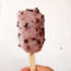 Image contains an off-white background with a frozen dessert on a stick in the foreground. The dessert is purplish in colour, and it contains numerous black and purple specks within it. Below the dessert, there is a wooden stick and a hand can be seen holding it.