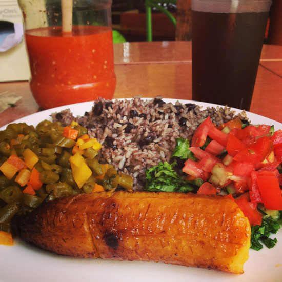 Image contains a photo of a plate of beans and rice with a small salad of tomato and lettuce on the right and a side of green beans and other chopped vegetables on the left. Below the beans and rice, there is a friend plantain. Above the plate, a bottle of chili sauce and a glass of a dark liquid can be seen.