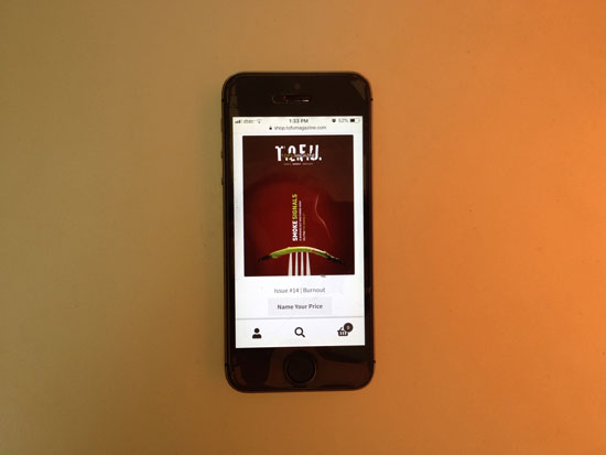 Image contains a photo of a small smartphone in the centre of the screen. On the smartphone, an online store is being viewed and there is a magazine cover with several buttons below it. The phone is laying on a background of faded orange.