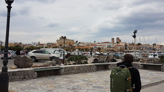 Image contains a photo of a man wearing a green backpack and a black hoodie. The man is in the bottom right of the photo, and he is facing away from the camera. In the distance, the horizon is dotted with small stone buildings and there are cars in the foreground.