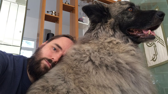 Image contains a photo of a bearded man with a large black and grey dog. The dog is sitting on its hind legs, and the man is resting his head on the dog's back just below his neck.
