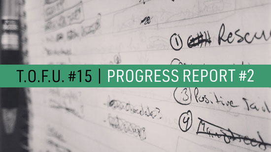 Image contains a notepad with illegible text in black ink and a pen on the left-hand side. In the foreground, there is text in the middle of the screen on a strip of light green that says "T.O.F.U. #15 |" in black and "Progress Report #2" in white.