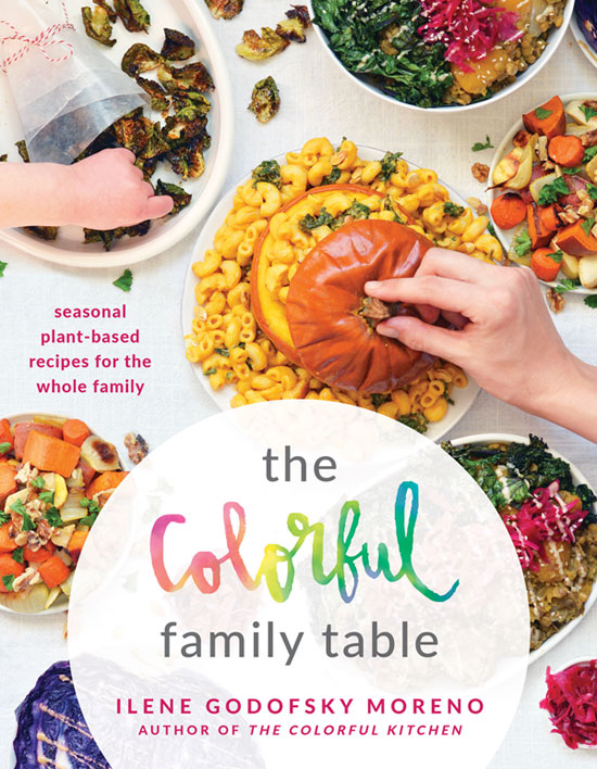 Image contains a variety of colourful food on a white table, including a roasted orange squash with macaroni, blue cabbage, a couple salads, and more. To the left of the centre of the page, there is pink text that sys â€œseasonal plant-based recipes for the whole familyâ€. In the bottom of the image, there is a frosted white circle with the words "The Colorful Family Table Ilene Godofsky Moreno Author of The Colorful Kitchen" in various colours.