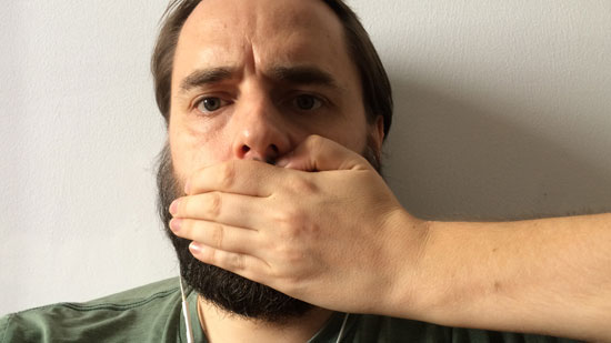 Photo contains a man with a beard looking directly at the camera. From the right-hand side, a hand and arm can be seen, and the hand is covering the man's mouth.