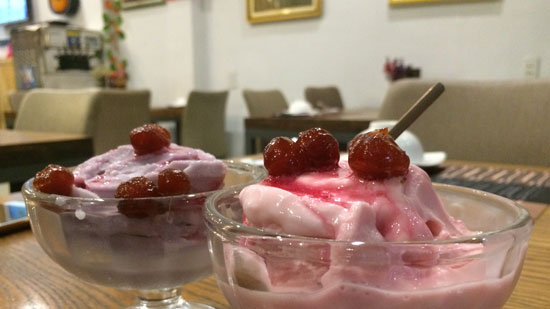 Image contains a photo of two small bowls of vegan ice cream on a restaurant table. The bowl in the foreground is filled with strawberry ice cream, which is pink. There are dark red berries on top of the ice cream along with a spoon. In the background, there is a bowl filled with a light purple ice cream, dark red berries, and a silver spoon.