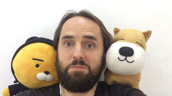 Image contains a photo of a man looking into the camera with an amused look on his face. On both shoulders, he has a stuffed animal.