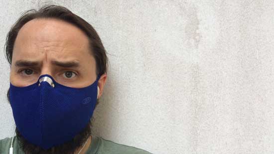 Image contains a photo of a man wearing a blue mask around the lower half of his face. The shoulders of the man are visible, and he's standing against a wall that is mostly white.