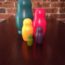 Image contains a photo of five wooden nesting dolls on a dark wooden table with a red brick background. From largest to smallest, the dolls are painted dark green, red, yellow, purple, and light green.
