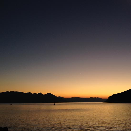 Image contains a photo of a sunset over a large body of water. On the horizon, hills can be seen stretching across most of the frame. The sky is a bright orange near the horizon, and it eventually turns a dark blue near the top of the frame.