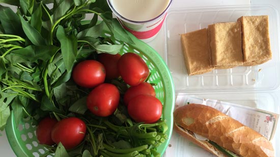 Image contains a photo of a variety of food on a white tabletop. On the left-hand side, in a green plastic basket, there are bright red tomatoes and a bunch of morning glory. To the right of the basket, there is a plastic container of small blocks of fried tofu, and just below that there is a banh mi sandwich.