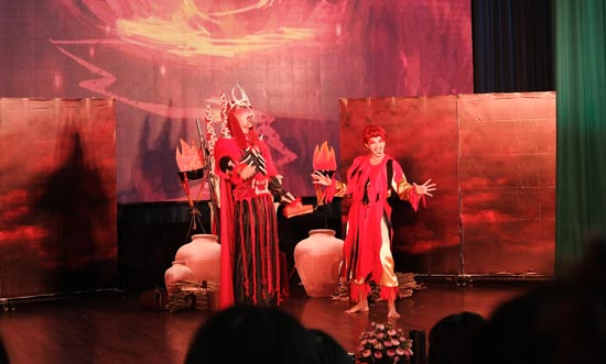 Image contains a photo of a theatrical production involving two characters in a setting similar to Hell. the male character is taller and wears a crown and armour, as well as a cape. The female character is striking a comedic pose with a smile on her face. Much of the photo is in red tones.