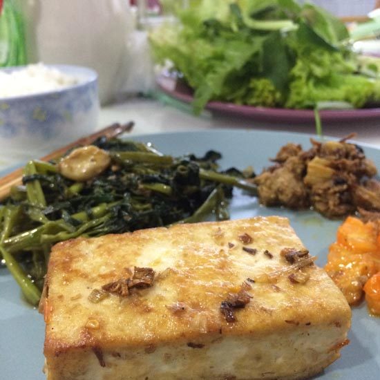 Image contains a photo of a block of fried tofu in the foreground. Behind it there are fried greens, kimchi, and lettuce leafs.