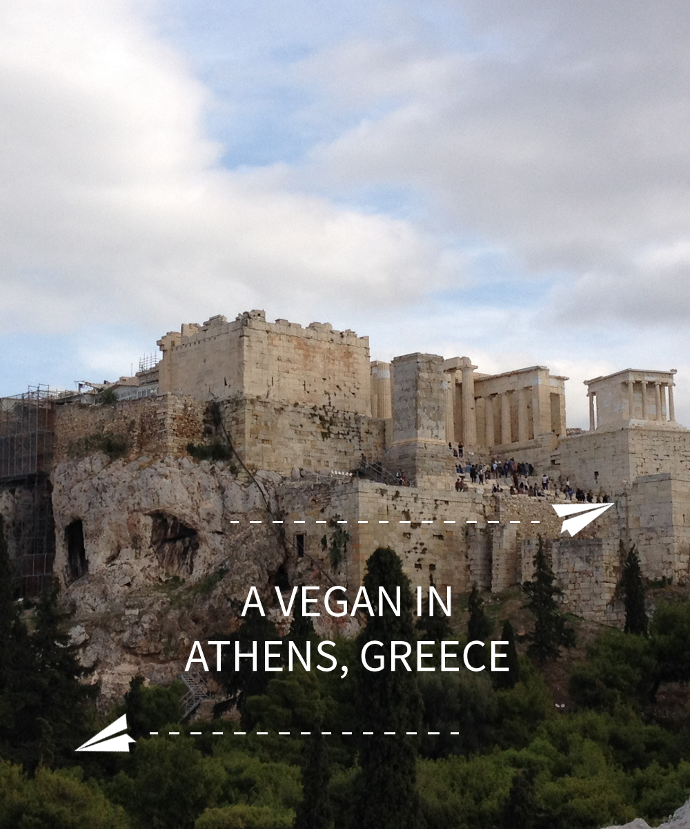 Image contains a photo of the Acropolis of Athens, Greece. Below the Acropolis, there is white text that says "A Vegan In Athens, Greece. Above the text, there is a illustration of a small paper plane with a dotted line trailing behind it. On the bottom left-hand side of the text, there is a similar illustration.