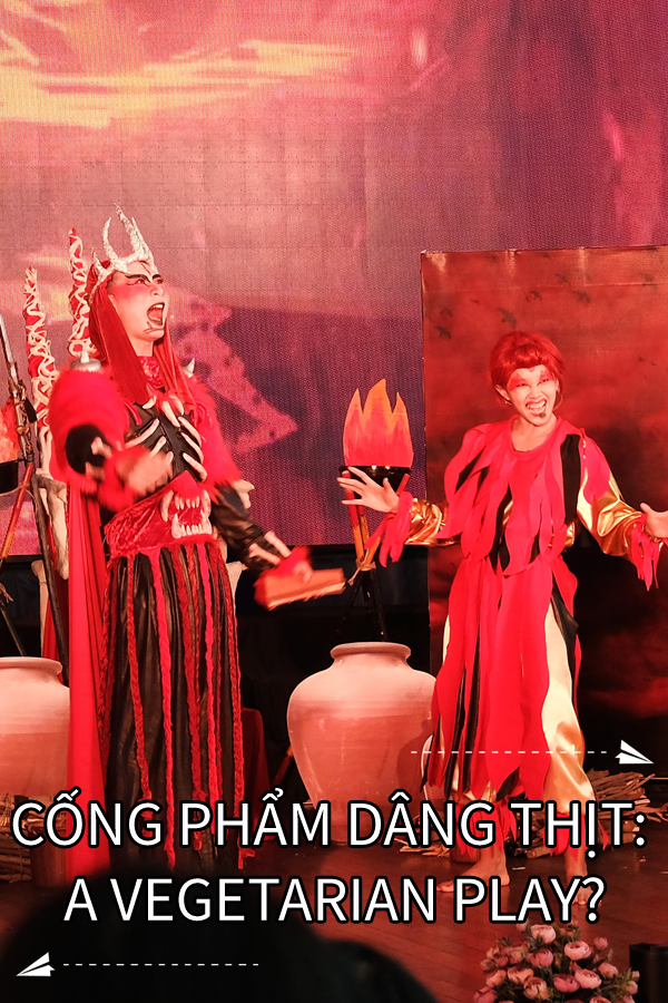 Image contains a photo of a theatrical production involving two characters in a setting similar to Hell. the male character is taller and wears a crown and armour, as well as a cape. The female character is striking a comedic pose with a smile on her face. Much of the photo is in red tones. In white text at the bottom of the image, there is a title that says "Cá»‘ng Pháº©m DÃ¢ng Thá»‹t: A Vegetarian Play?". On the right-hand side above the text, there is a small white paper airplane with a dotted line behind it. Below the text and on the left-hand side, there is a similar icon.