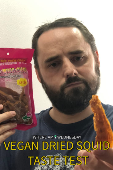Image contains a photo of a bearded man holding a dried piece of food in his fingers in the foreground of the shot. The man is facing the camera, and he is holding a red plastic pouch of a Vietnamese food product in his right hand. In the bottom of the image, there is small white text that says "Where Am I Wednesday". Below that, there is larger yellow text that says "Vegan Dried Squid Taste Test".