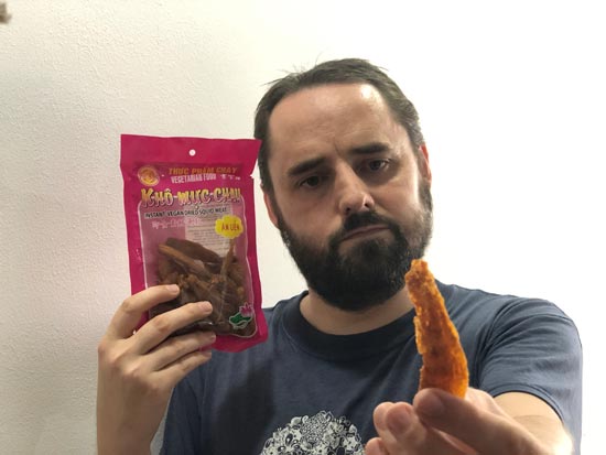 Image contains a photo of a bearded man holding a dried piece of food in his fingers in the foreground of the shot. The man is facing the camera, and he is holding a red plastic pouch of a Vietnamese food product in his right hand.