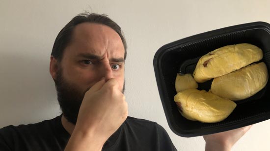 Image contains a photo of a bearded white man pinching his nose and looking unhappy. To the right of the man, there is a black plastic tray of durian, which is a yellowish fruit shaped in large pieces.