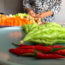 Image contains a photo of a number of vegetables. In the foreground, there are red chilis and green bitter melons of various sizes. In the background, there are cubed carrots, mushrooms, and more. A woman's hands can be seen near the vegetables in the background.
