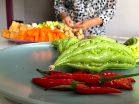 Image contains a photo of a number of vegetables. In the foreground, there are red chilis and green bitter melons of various sizes. In the background, there are cubed carrots, mushrooms, and more. A woman's hands can be seen near the vegetables in the background.