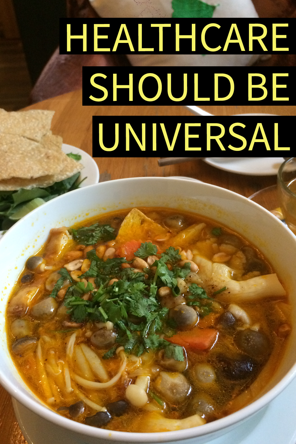 Image contains a photo of a white bowl of soup and noodles in the lower two-thirds of the frame, and a glass cup of tea in the top right-hand corner. In the foreground above the tea cup, there is yellow text on the left-hand side that says "Healthcare Should Be Universal" on top of black bars.