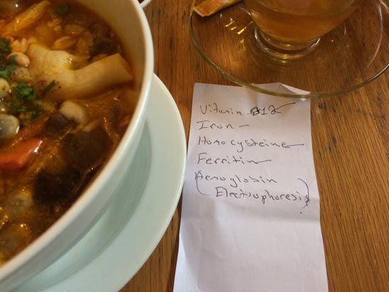 Image contains a photo of a white bowl of soup and noodles on the left, a glass cup of tea in the top right-hand corner, and a paper note in the bottom right-hand. On the note, there is handwriting that says "Vitamin B12, iron, homocysteine, ferritin, and hemoglobin electrophoresis."