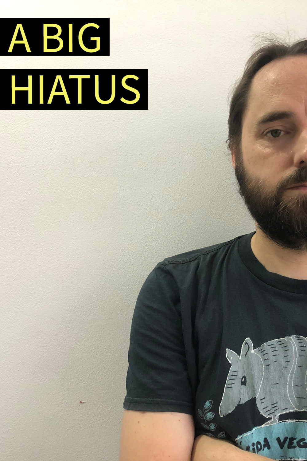 Image contains a photo of a bearded white man in a tshirt with his arms crossed. He is placed to the right of the photo and is looking directly at the camera. To the left, there is yellow text on black blocks that says "A Big Hiatus".