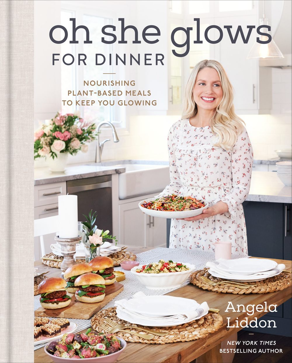 Image contains a photo of a white woman holding a plate of food in a kitchen. In front of her, there is a wooden table with a number of other dishes. To the left and above her, there is text that says "Oh She Glows for Dinner" and "Nourishing Plant-Based Meals to Keep You Glowing". In the bottom right-hand side, there is white text that says "Angela Liddon."